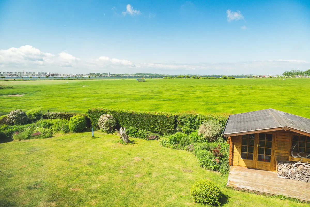 Der Ausblick aus der OG 2 im Sommer: unten der Garten mit Hecke und Holzschuppen, dahinter weite grüne Felder bis zum Binnensee, am Horizont Häuser und Bäume unter blauem Himmel