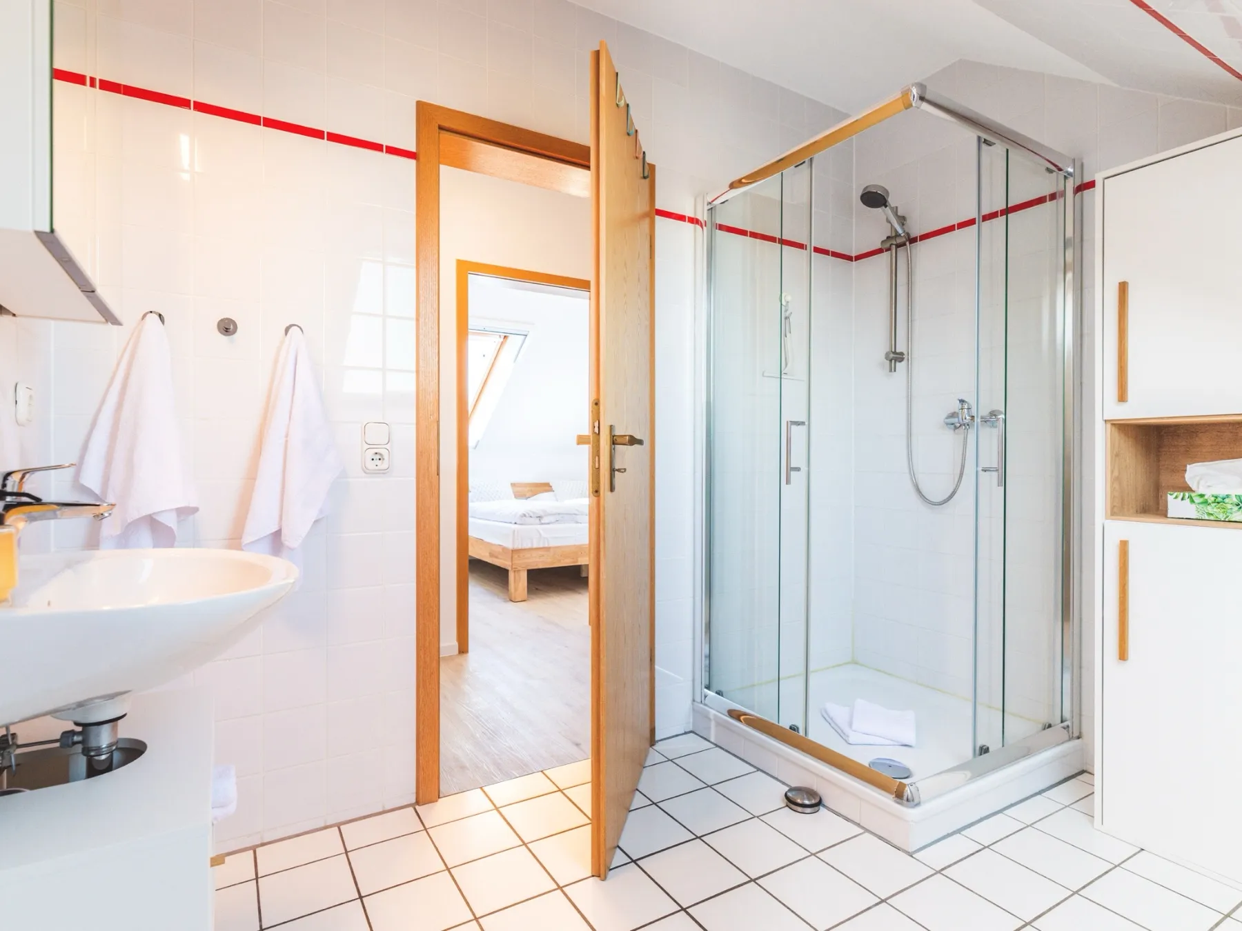 Das Bad aus der zweiten Blickrichtung: rechts die gläserne Eckdusche mit Handbrause, links das Waschbecken mit Spiegelschrank und zwei Handtüchern an der Wand, weiße Fliesen mit schmalem rotem Fliesenband, in der Mitte die offene Tür mit Blick auf das Bett im Schlafzimmer