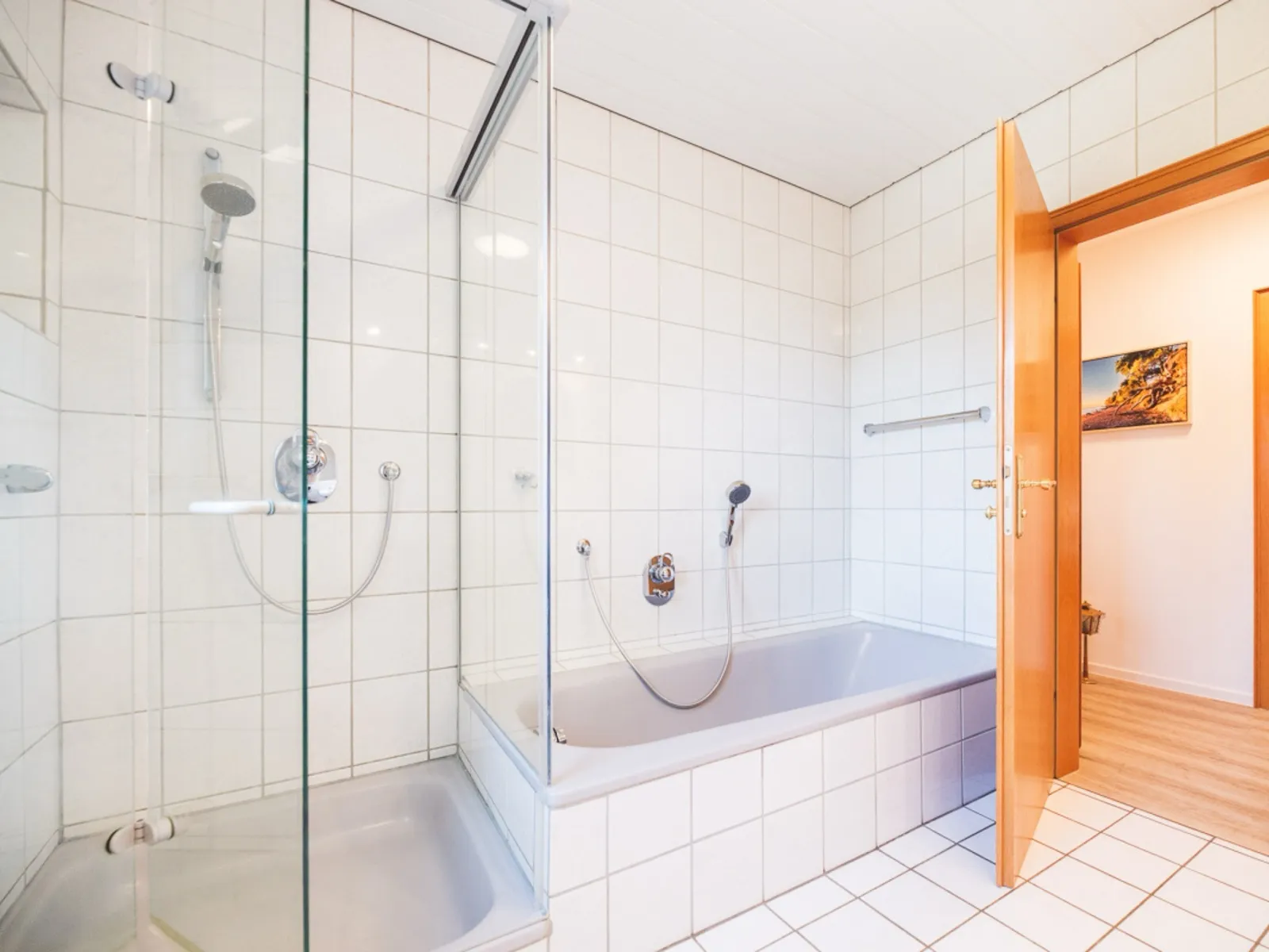 Zweite Ansicht desselben Badezimmers: links die gläserne Eckdusche mit Handbrause, rechts daneben die eingemauerte Badewanne, ganz rechts die geöffnete Holztür mit Blick in den Flur