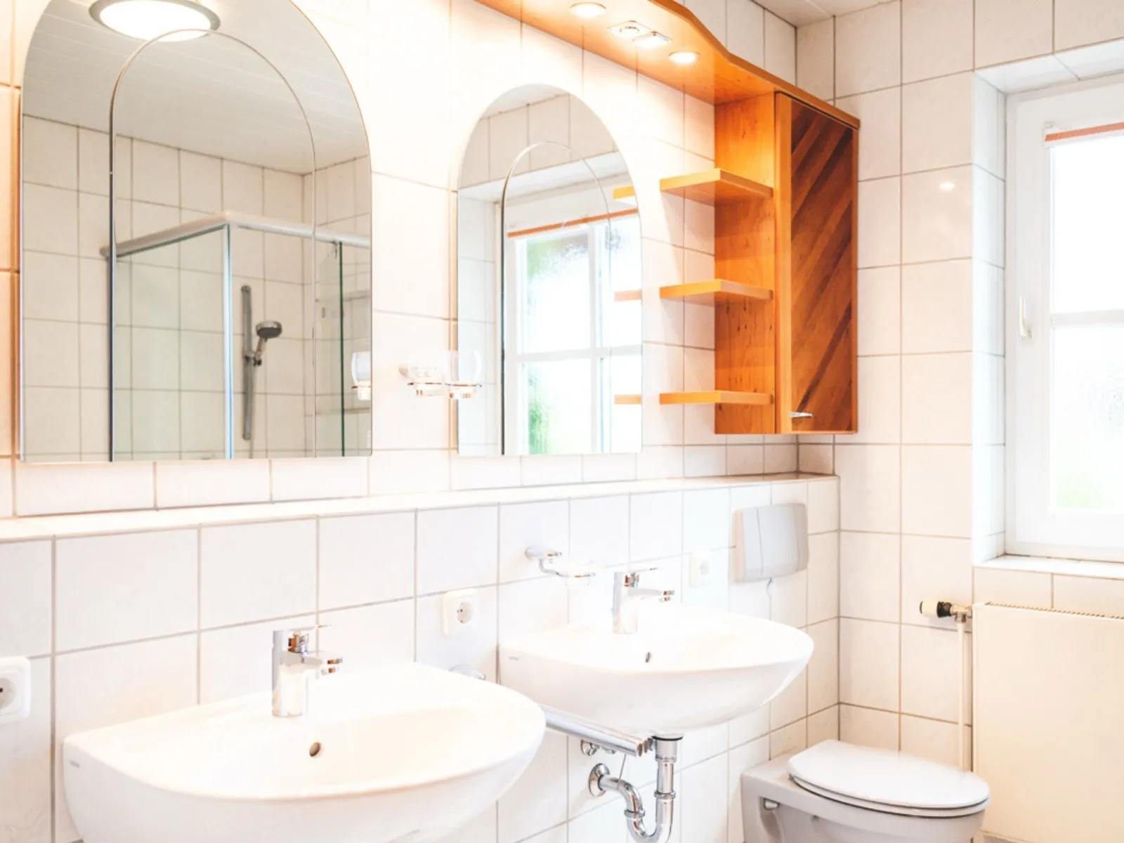 Das große Badezimmer: zwei Waschbecken nebeneinander, darüber je ein hoher Bogenspiegel, rechts ein Hängeschrank aus Holz mit offenen Ablagen, daneben das WC und ein Fenster, durch das Tageslicht hereinfällt