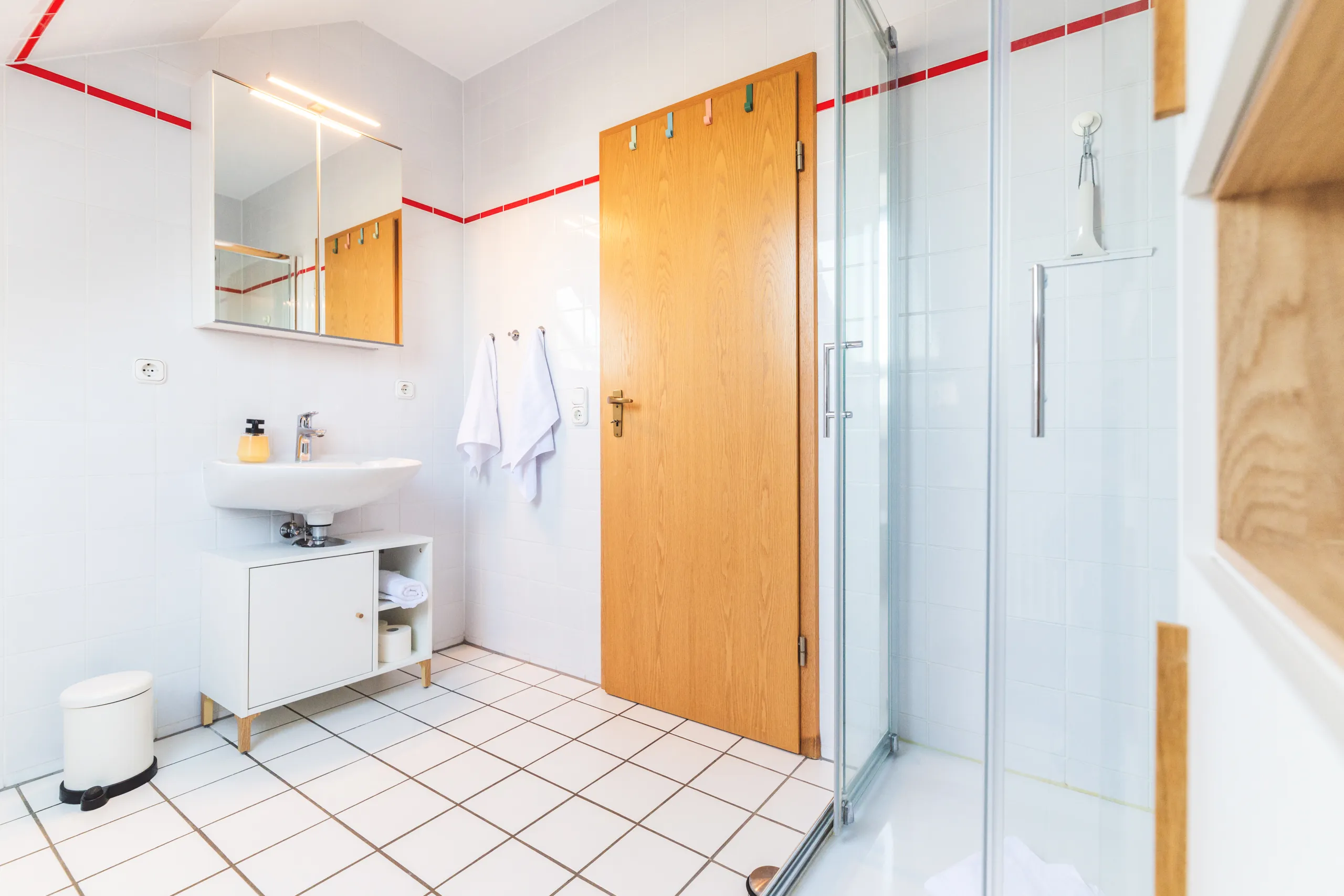 Badezimmer mit Glasdusche, Waschbecken mit Unterschrank und Spiegelschrank, weiß gefliest mit schmalem rotem Fliesenband
