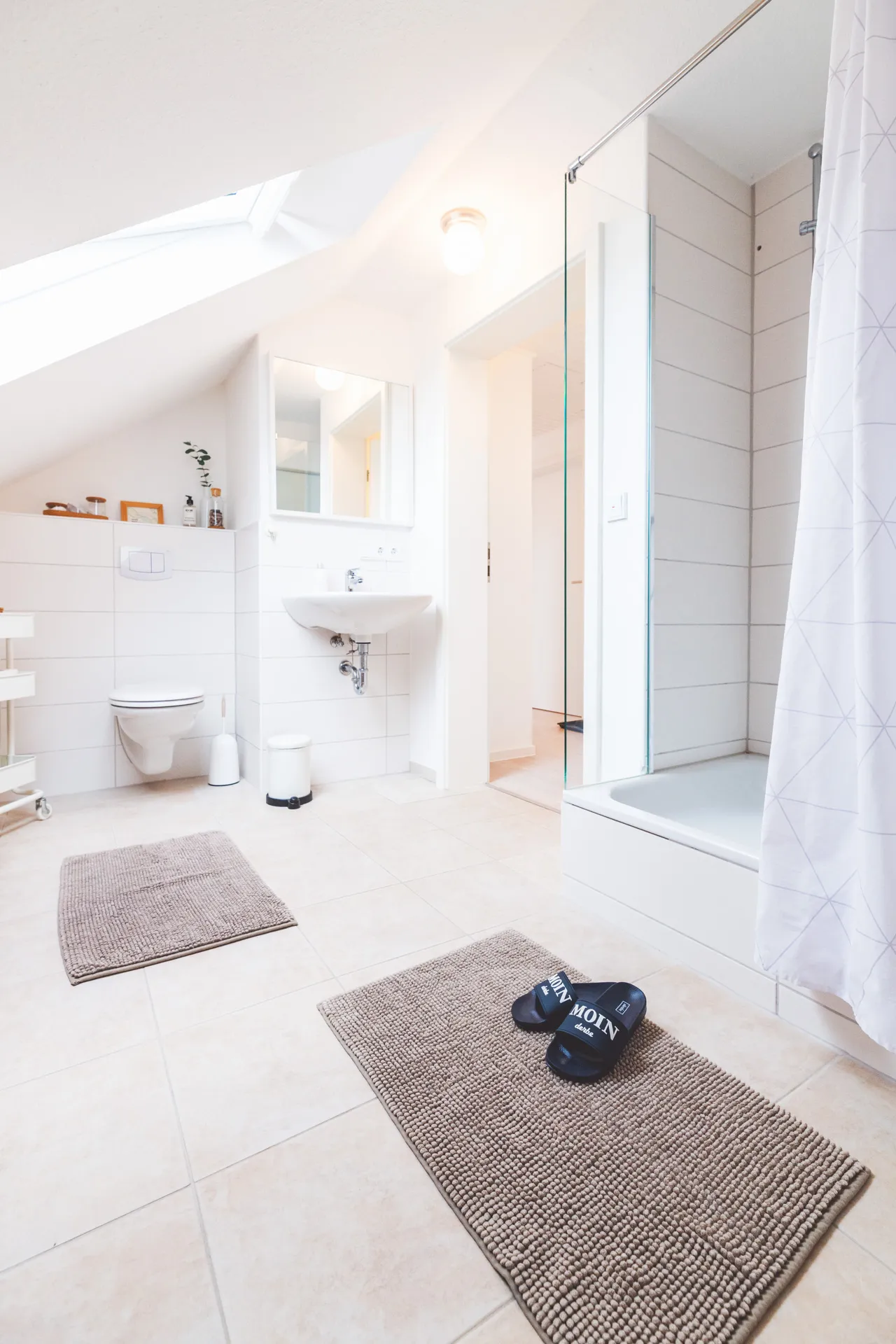 Bad mit Wand-WC, Waschbecken und Spiegelschrank, rechts die Dusche hinter einer Glaswand, auf den hellen Fliesen zwei Badvorleger