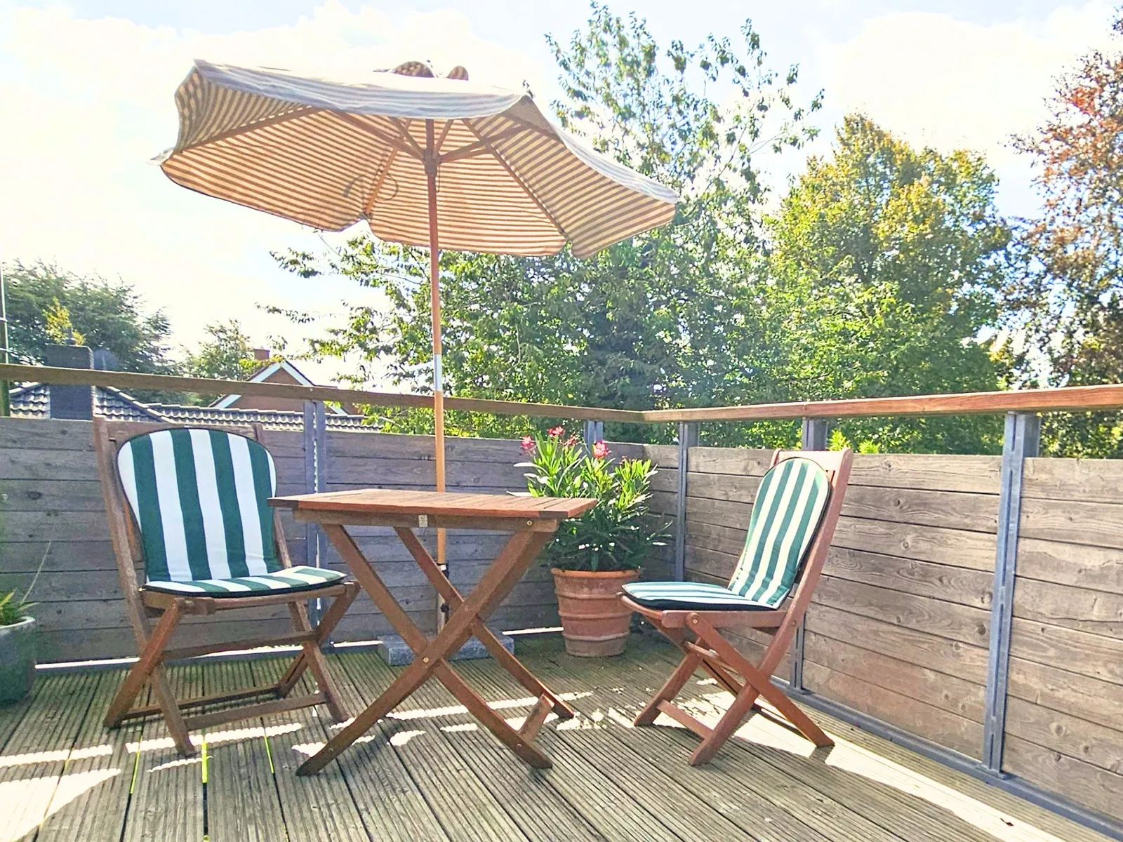 Der Balkon mit aufgespanntem, beige-weiß gestreiftem Sonnenschirm über einem Holz-Klapptisch mit zwei Klappstühlen mit grün-weiß gestreiften Auflagen, daneben ein blühender Oleander im Terrakottatopf, dahinter Holzsichtschutz und Bäume in der Sonne