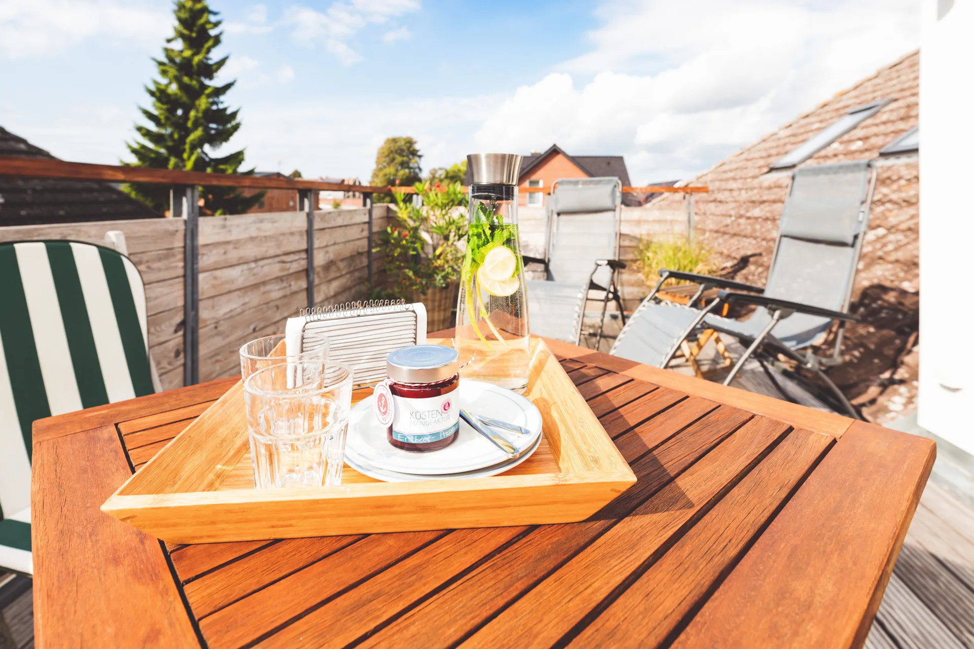 Holztablett auf dem Balkontisch mit Wasserkaraffe, zwei Gläsern und einem Glas Erdbeermarmelade, dahinter die Relaxliegen
