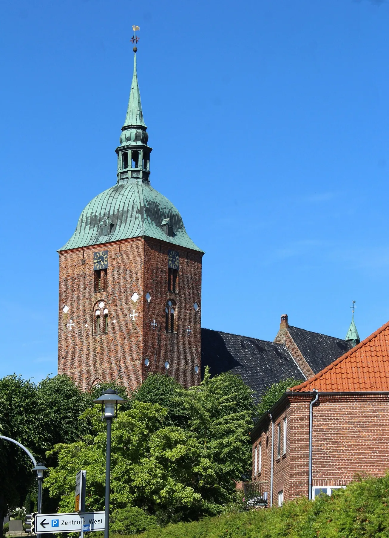 Der Turm der St.-Nikolai-Kirche in Burg: rotes Backsteinmauerwerk mit einer grün verwitterten Kupferhaube, daneben Bäume und ein Ziegeldach.