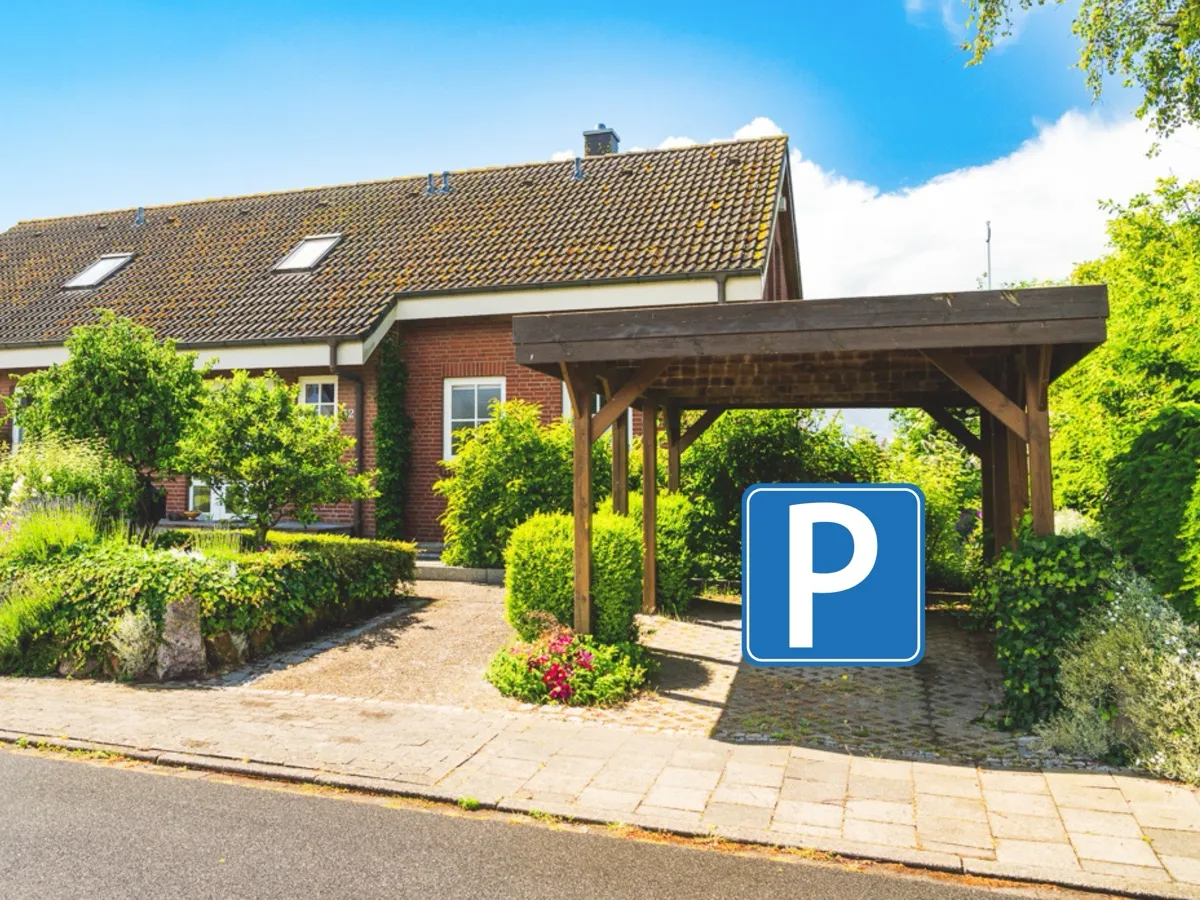 Das Huus Binnensee von der Straße aus: rotes Klinkerhaus mit Dachfenstern hinter niedrigen Hecken und blühenden Stauden, rechts der dunkle Holz-Carport über der gepflasterten Auffahrt; darin markiert ein großes blaues Parkplatz-Zeichen den Stellplatz.