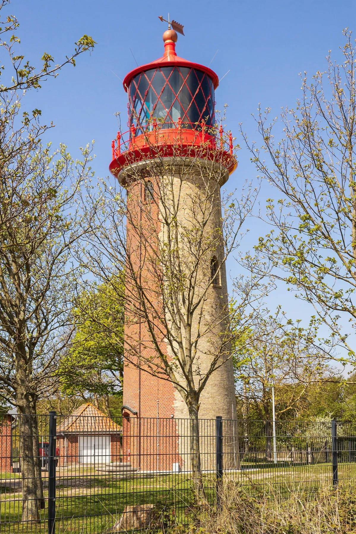 Der Leuchtturm Staberhuk an der Ostspitze der Insel: ein runder Backsteinturm mit rotem Laternenhaus, umgeben von hellgrünen Frühlingsbäumen unter blauem Himmel.