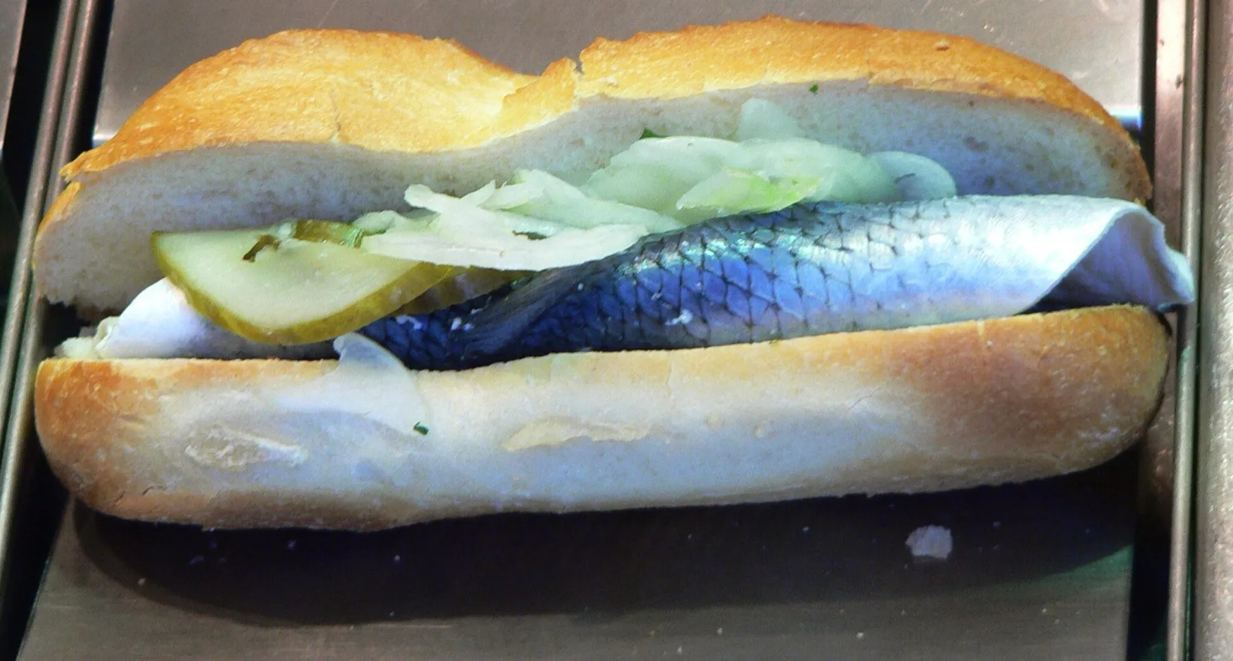 Ein aufgeklapptes Fischbrötchen mit einem Heringsfilet, Zwiebelringen und einer Scheibe Gewürzgurke.