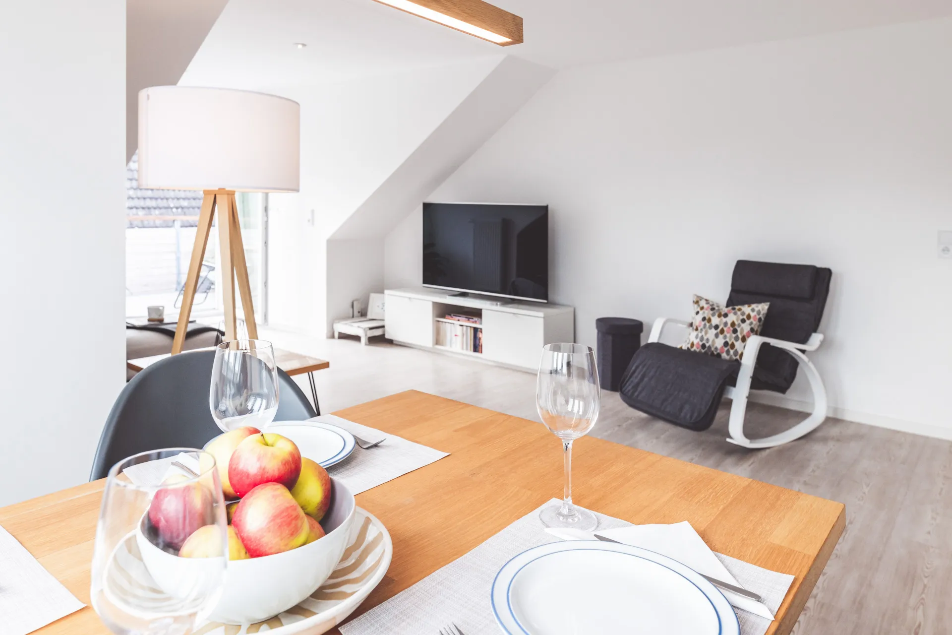 Gedeckter Esstisch mit Obstschale und Weingläsern, dahinter der Wohnbereich mit Smart-TV und Schaukelsessel