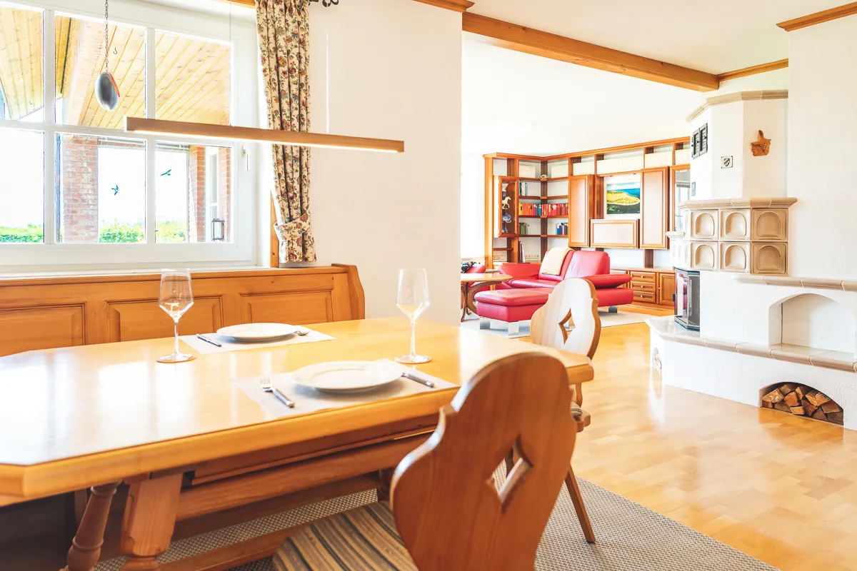 Esszimmer mit massivem Holztisch und geschnitzten Stühlen, im Hintergrund der Wohnbereich mit rotem Ledersofa und weißem Kachelofen