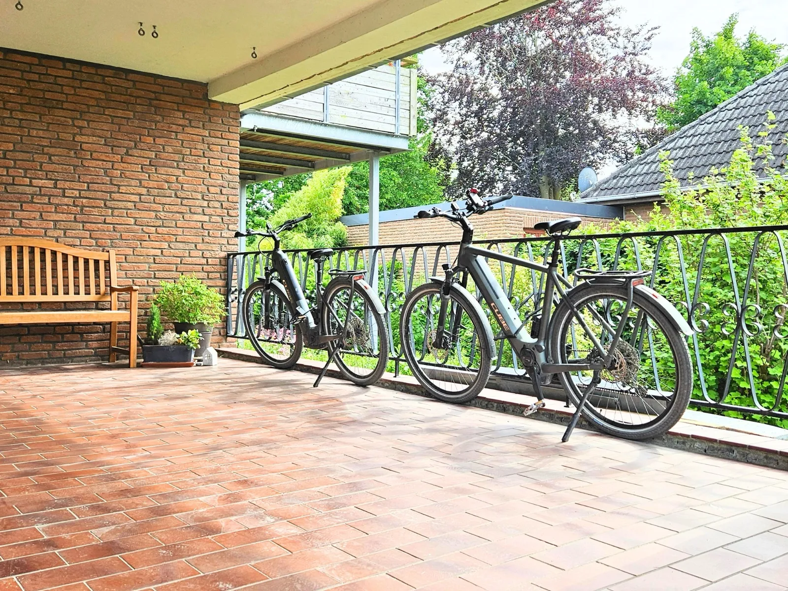 Zwei dunkle E-Bikes stehen vor dem Haus an einem schmiedeeisernen Metallgeländer, auf der gepflasterten Fläche unter dem vorstehenden Balkon; links eine Holzbank an der Backsteinwand mit Kräutertöpfen, rechts eine hohe Hecke