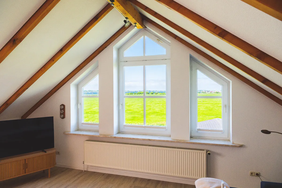 Die große Fensterfront im Giebel des Wohnzimmers: drei Fenster unter sichtbaren Dachbalken, das mittlere mit spitzem Abschluss, davor ein Heizkörper, links der Fernseher auf einem Sideboard; draußen der Blick über die Felder Richtung Binnensee