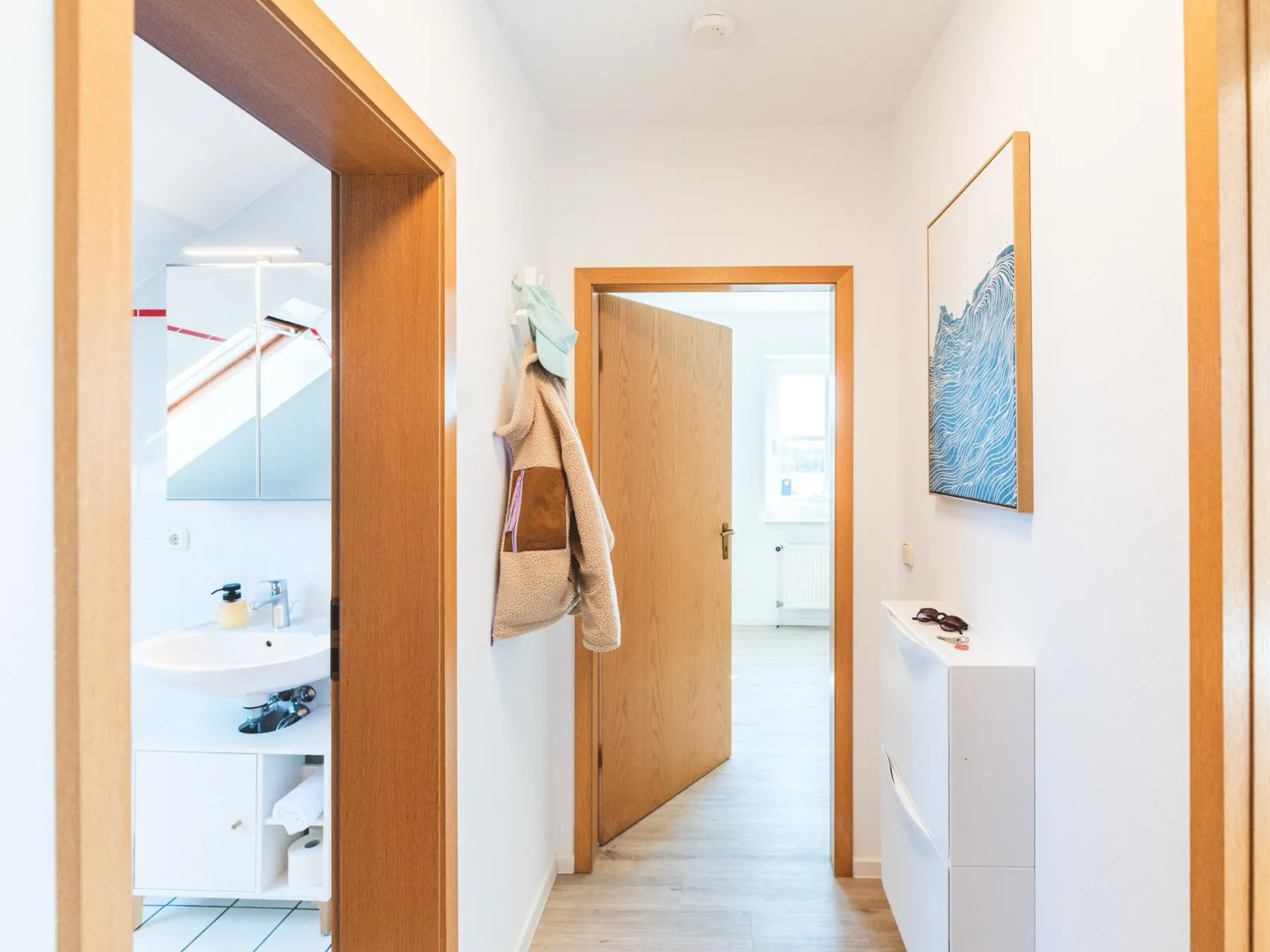 Der Flur der Wohnung: links die offene Badtür mit Waschbecken und Spiegelschrank, in der Mitte eine offene Zimmertür mit Blick in den hellen Wohnbereich, an der Wand ein Garderobenhaken mit Jacke und Mütze, rechts ein gerahmter blauer Wellen-Kunstdruck über einem schmalen weißen Schuhschrank