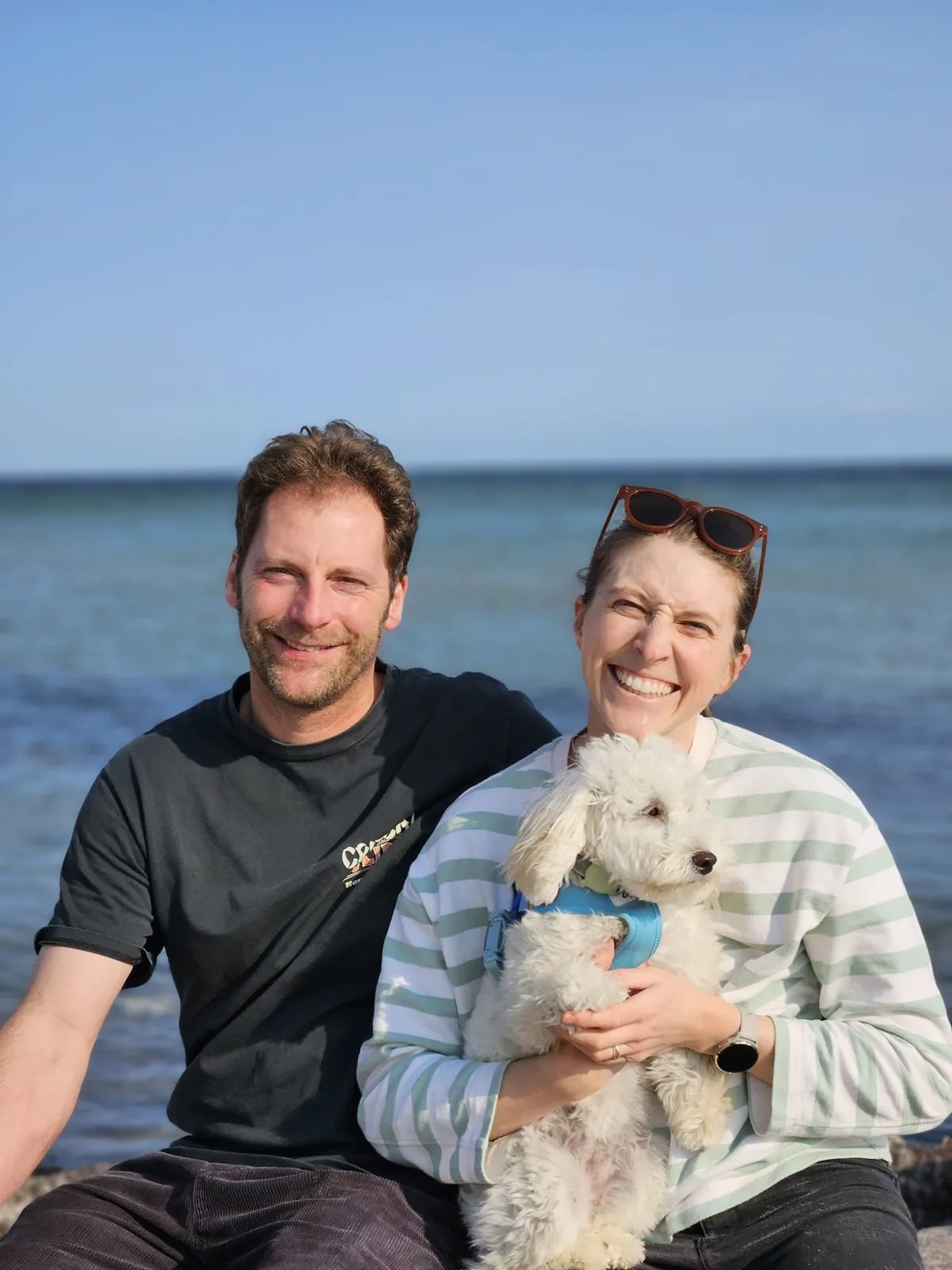 Friederike und Jan Ueberall sitzen dicht beieinander am Wasser und lachen in die Kamera; Friederike hält einen kleinen weißen Hund im Arm, hinter ihnen offenes Wasser unter blauem Himmel.