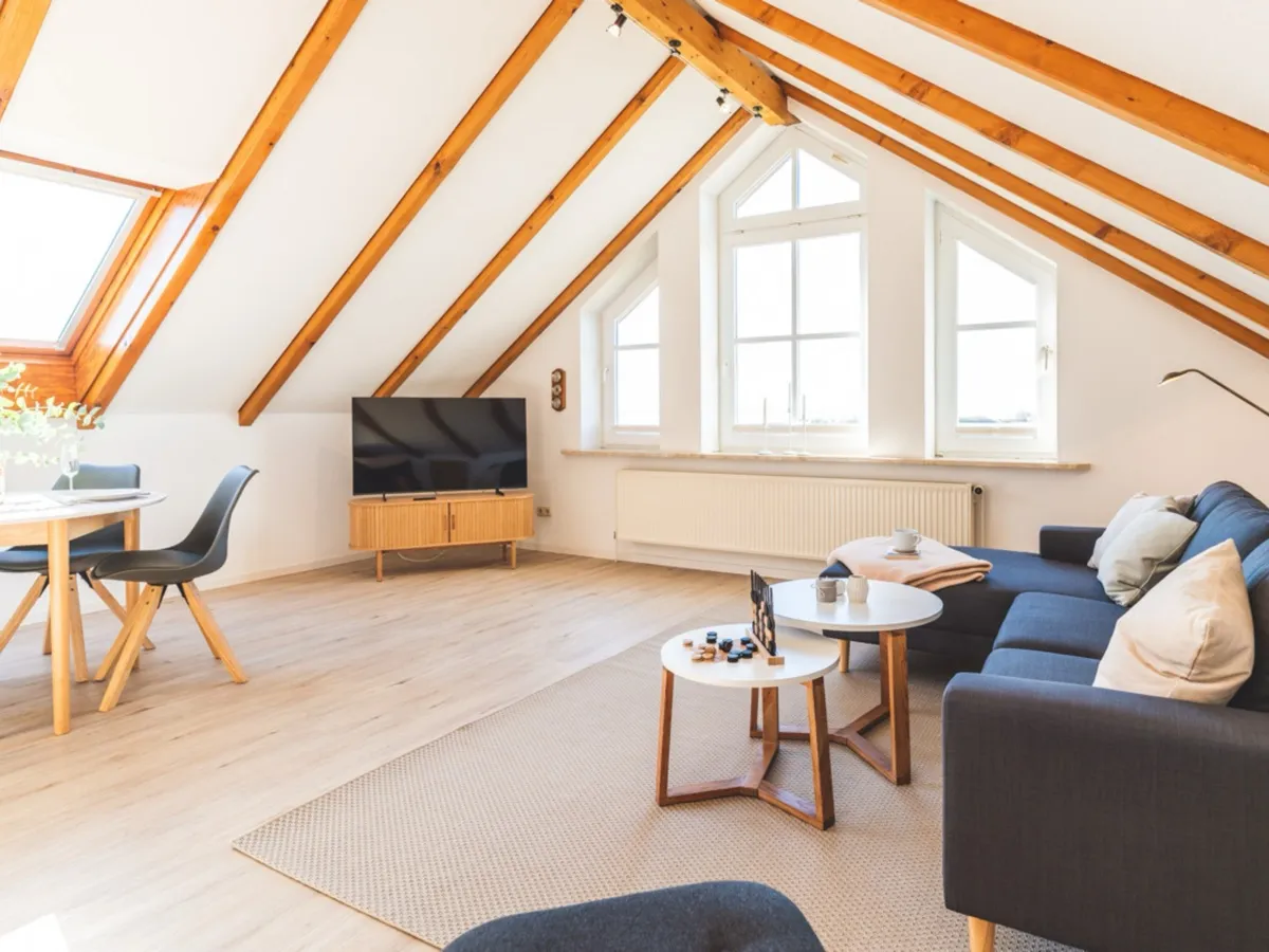 Der Wohnbereich Richtung Giebel: der Fernseher auf einem hellen Holz-Sideboard, dahinter die dreiteilige Fensterfront mit dem spitz zulaufenden Mittelfenster und einem Heizkörper unter der Fensterbank; rechts das blaue Sofa mit den beiden runden Beistelltischen, links der Esstisch, darüber die sichtbaren Dachsparren.