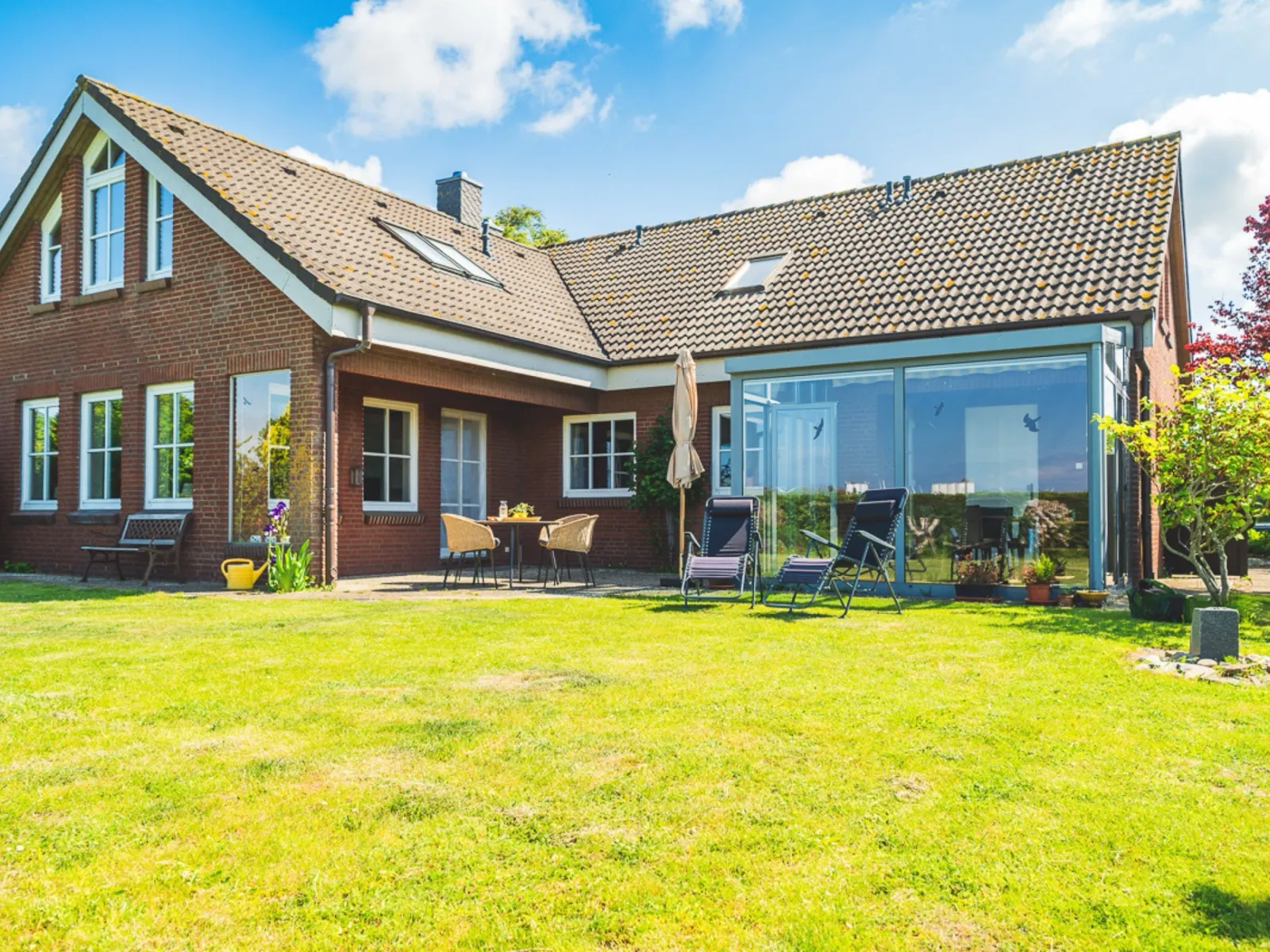 Das Backsteinhaus vom eigenen Garten aus: rechts der verglaste Wintergarten mit zwei Relaxliegen davor, in der Mitte die überdachte Terrasse mit Tisch und Korbstühlen, links eine Bank an der Hauswand, im Vordergrund der große Rasen unter blauem Himmel