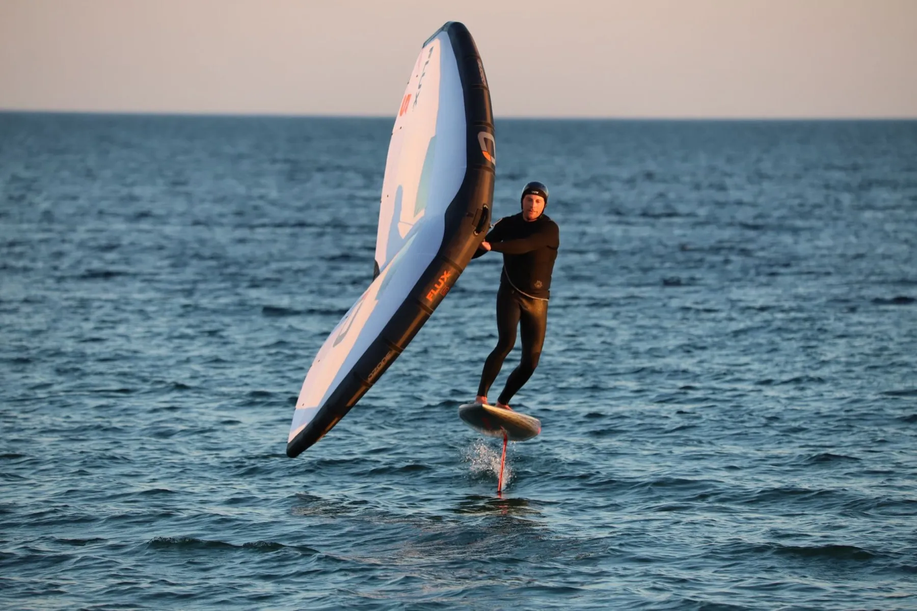 Wingfoilen auf dem Wasser: ein Sportler im dunklen Neoprenanzug hält den hellen Wing schräg über sich, das Board schwebt auf dem Foil über der Oberfläche, dahinter offenes Wasser im warmen Abendlicht.