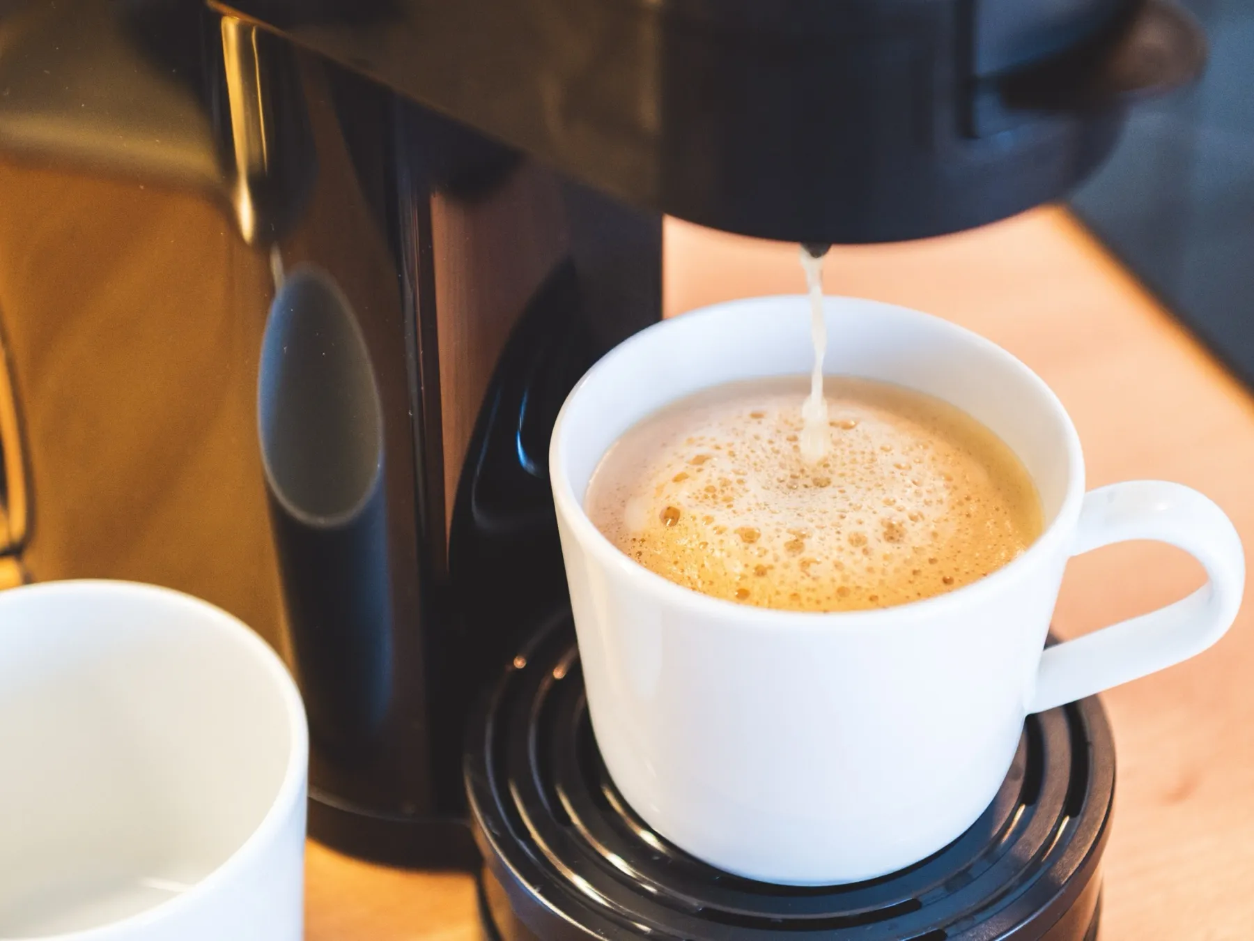 Aus der schwarzen Kaffeemaschine auf der hellen Holzarbeitsplatte läuft Kaffee mit feiner Crema in eine weiße Tasse, links im Anschnitt steht eine zweite Tasse bereit