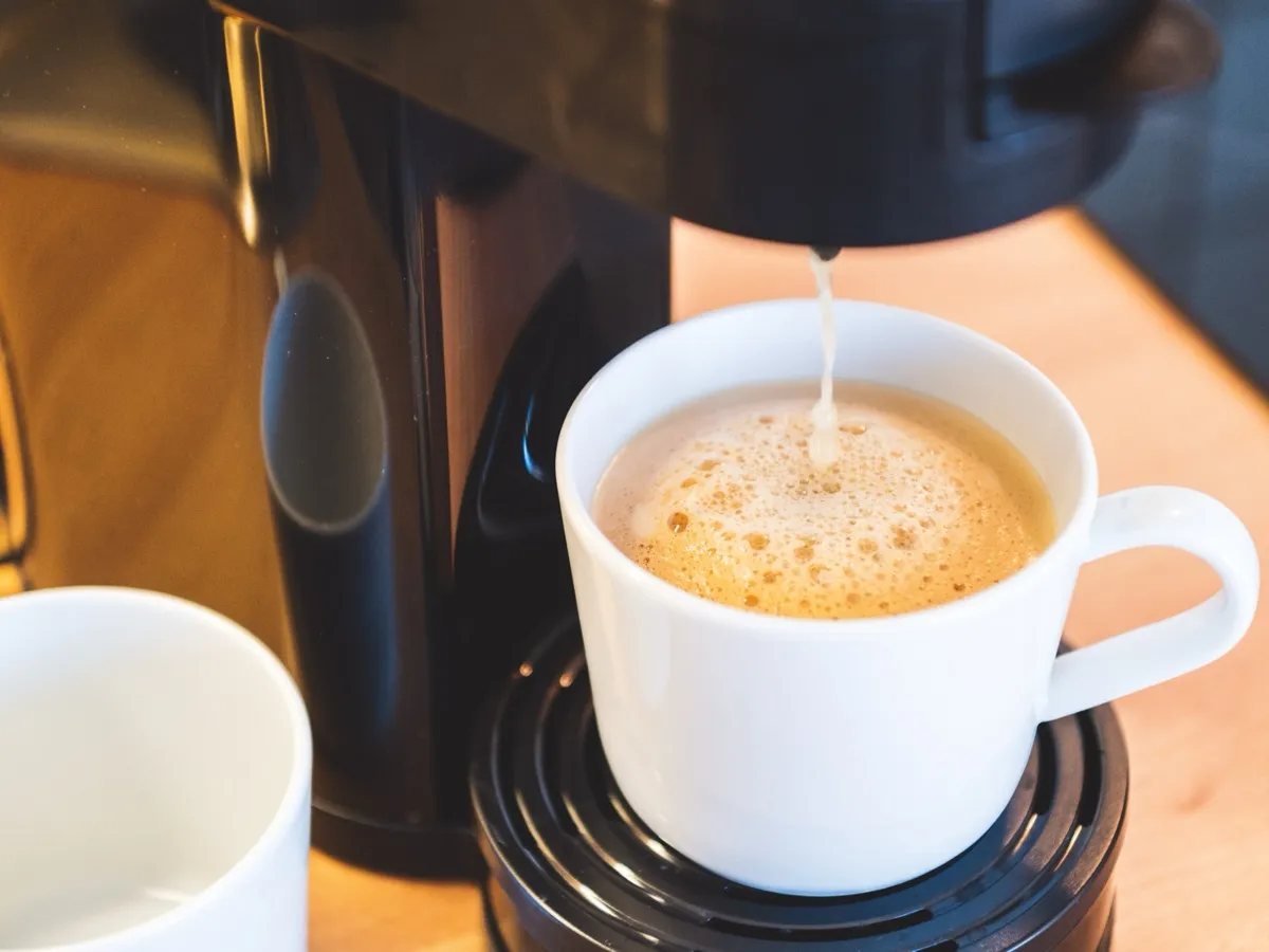 Frisch gebrühter Kaffee läuft aus der schwarzen Kaffeemaschine in eine weiße Tasse mit Crema, links daneben steht eine zweite Tasse bereit; darunter die helle Holzarbeitsplatte der Pantryküche.