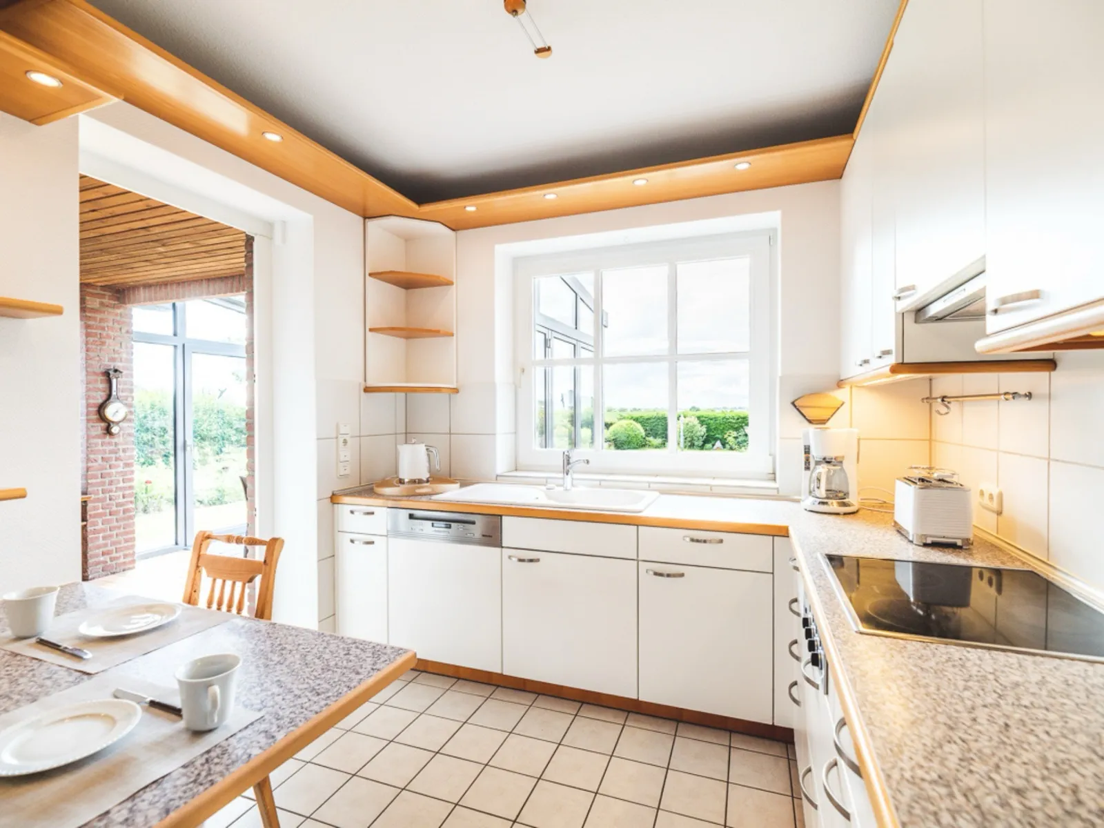 Helle Küche mit weißer Zeile und Holzkanten: über der Spüle ein Fenster mit Blick auf Hecke und Garten, darunter die integrierte Spülmaschine, daneben Wasserkocher, Kaffeemaschine und Toaster, rechts das Ceranfeld mit Dunstabzug; links führt ein Durchgang an der Backsteinwand vorbei in den Wintergarten, vorne links steht der gedeckte Frühstückstisch