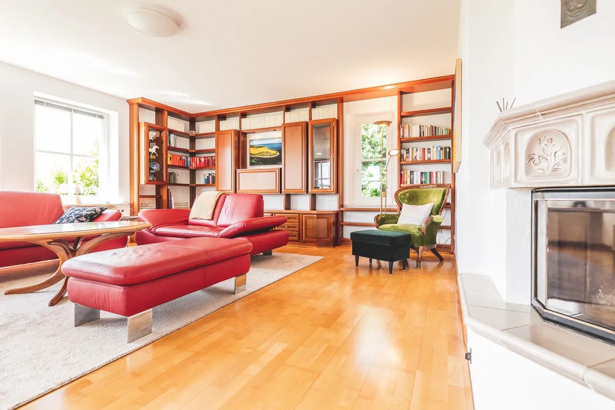 Wohnbereich mit rotem Ledersofa und Hocker, grünem Ohrensessel, einer Bücherwand aus Holz und dem Kachelofen mit Sichtfenster