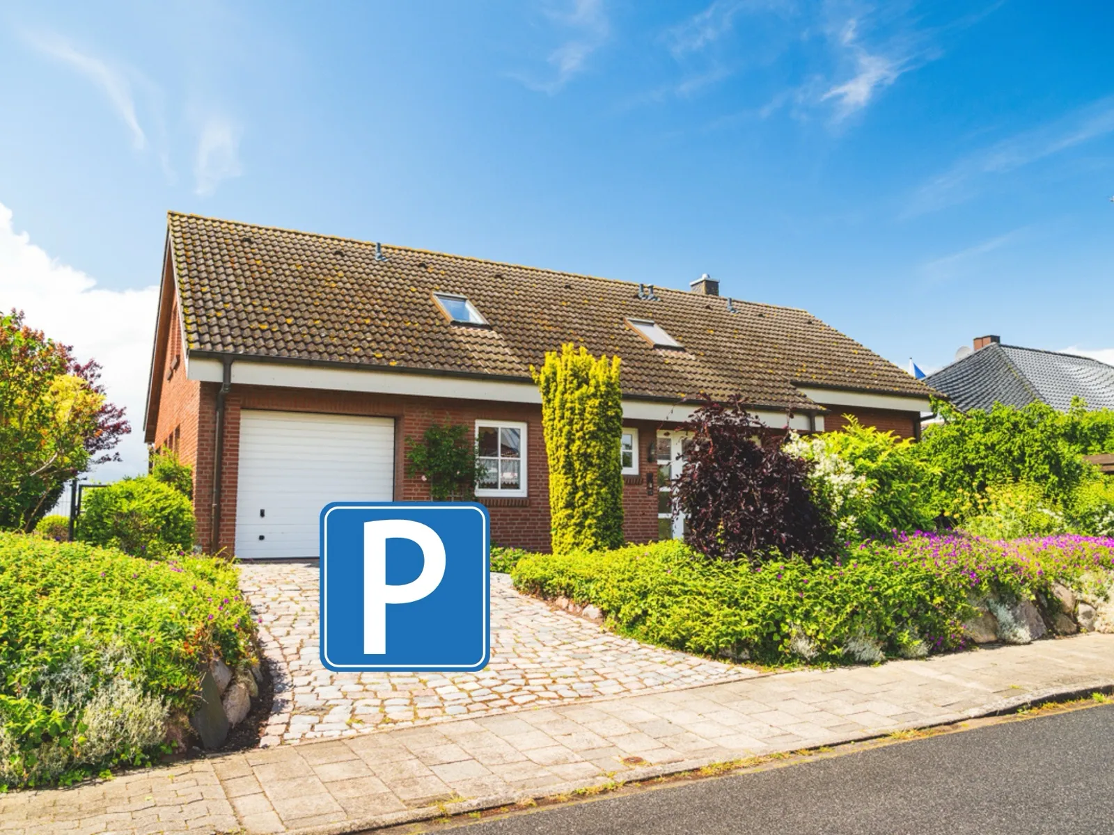 Das Haus von der Straße aus mit weißem Garagentor und gepflasterter Auffahrt, umgeben von Hecken und blühenden Stauden; ein blaues Parkplatz-Symbol im Bild markiert den Stellplatz vor der Garage