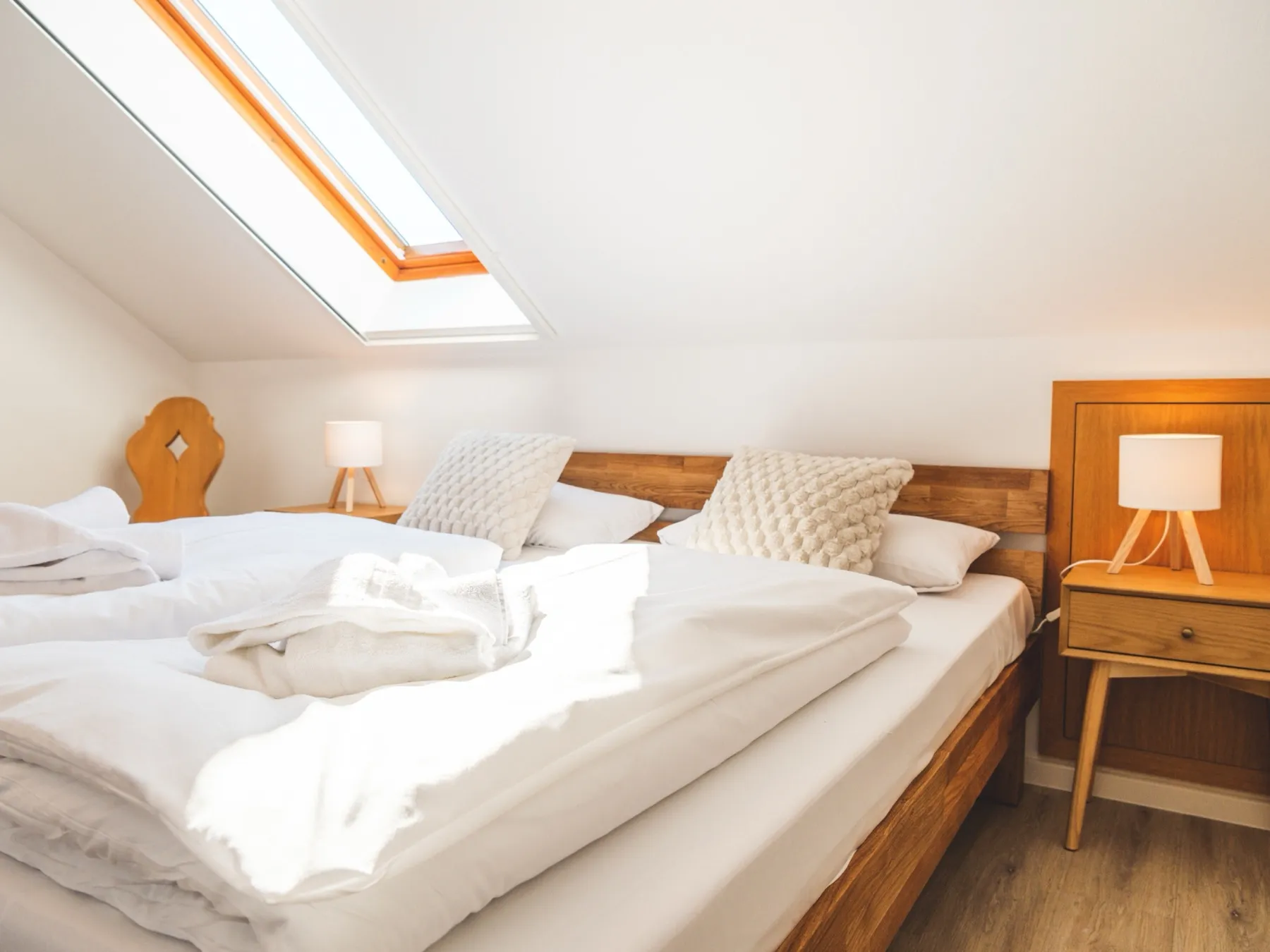 Dasselbe Schlafzimmer von der Fußseite aus: das Doppelbett längs im Raum vor dem hölzernen Kopfteil, rechts ein Nachttisch aus Eiche mit Tischleuchte, links unter der Dachschräge das geöffnete Dachfenster und ein Stuhl mit geschnitzter Holzlehne