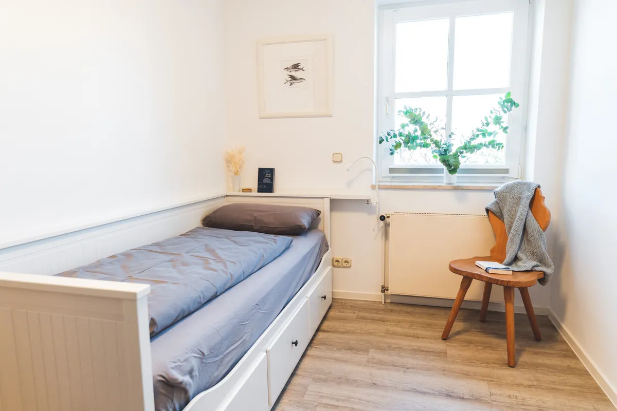 Kleines zweites Schlafzimmer mit weißem Einzelbett, ausziehbar auf 160 × 200 cm, daneben ein Holzhocker am Fenster