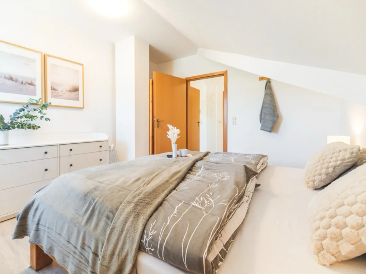 Das Schlafzimmer von der Kommodenseite: das Doppelbett mit graubrauner Tagesdecke und beigen Strukturkissen unter der Dachschräge, links eine weiße Kommode mit Eukalyptus in der Vase und zwei gerahmten Dünenbildern darüber, dahinter die offene Tür zum Bad, rechts ein Handtuch am Haken.