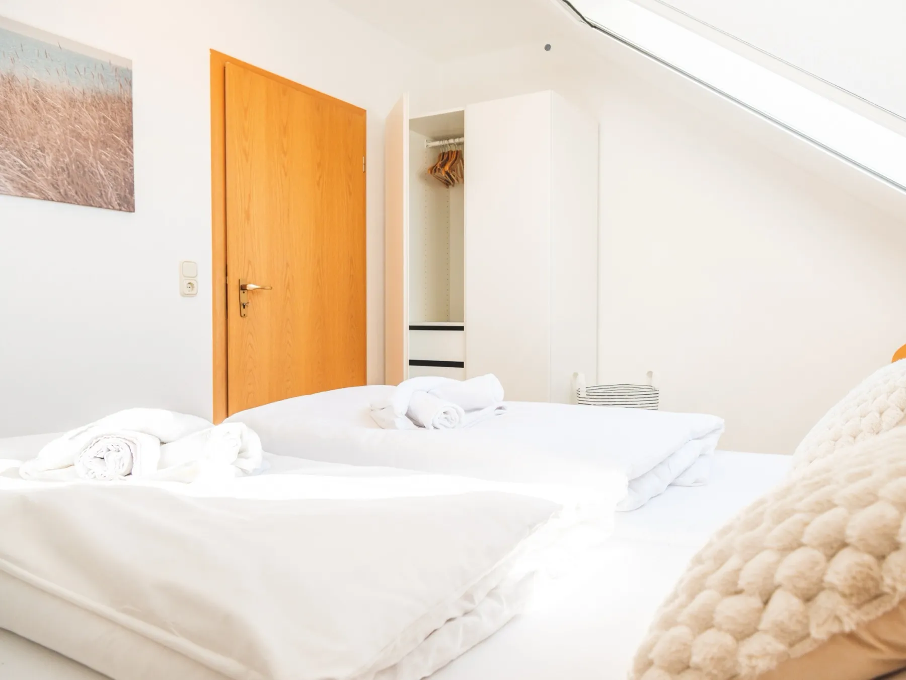 Blick vom Bett zur Zimmertür: ein offener weißer Kleiderschrank mit Kleiderstange, Bügeln und Schubladen, daneben die Zimmertür aus Eiche, links an der Wand ein gerahmtes Foto von Strandhafer, rechts die Dachschräge mit dem Fenster