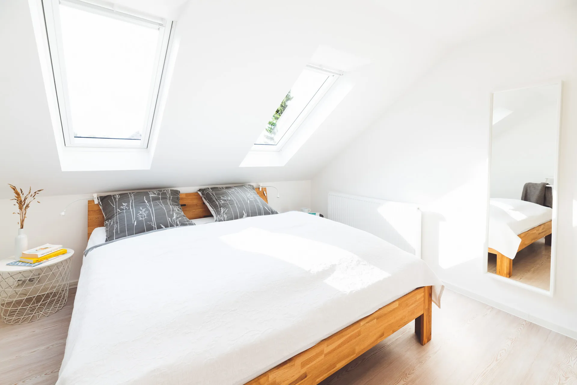 Schlafzimmer unter der Dachschräge mit Doppelbett aus Eiche und weißer Tagesdecke, zwei Dachfenster, rechts ein hoher Standspiegel
