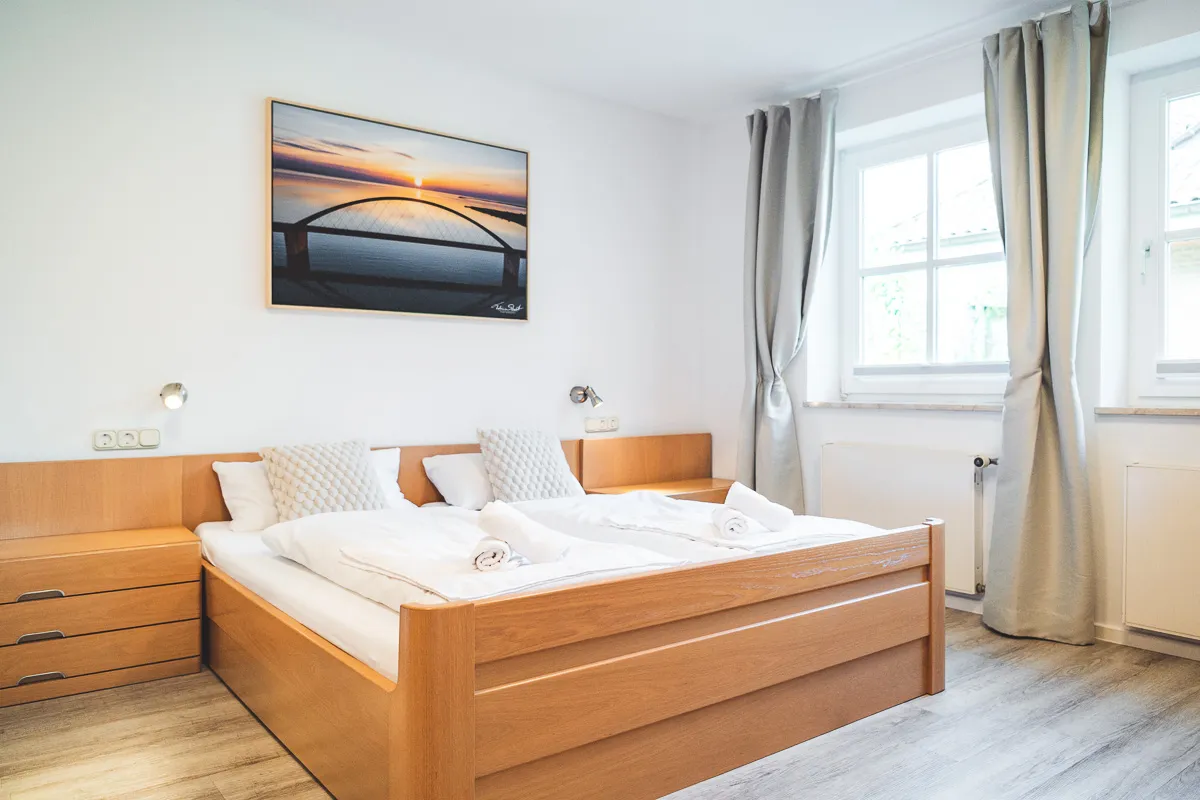 Schlafzimmer mit Doppelbett aus Eiche und frisch bezogener Bettwäsche, darüber ein Foto der Fehmarnsundbrücke im Sonnenuntergang