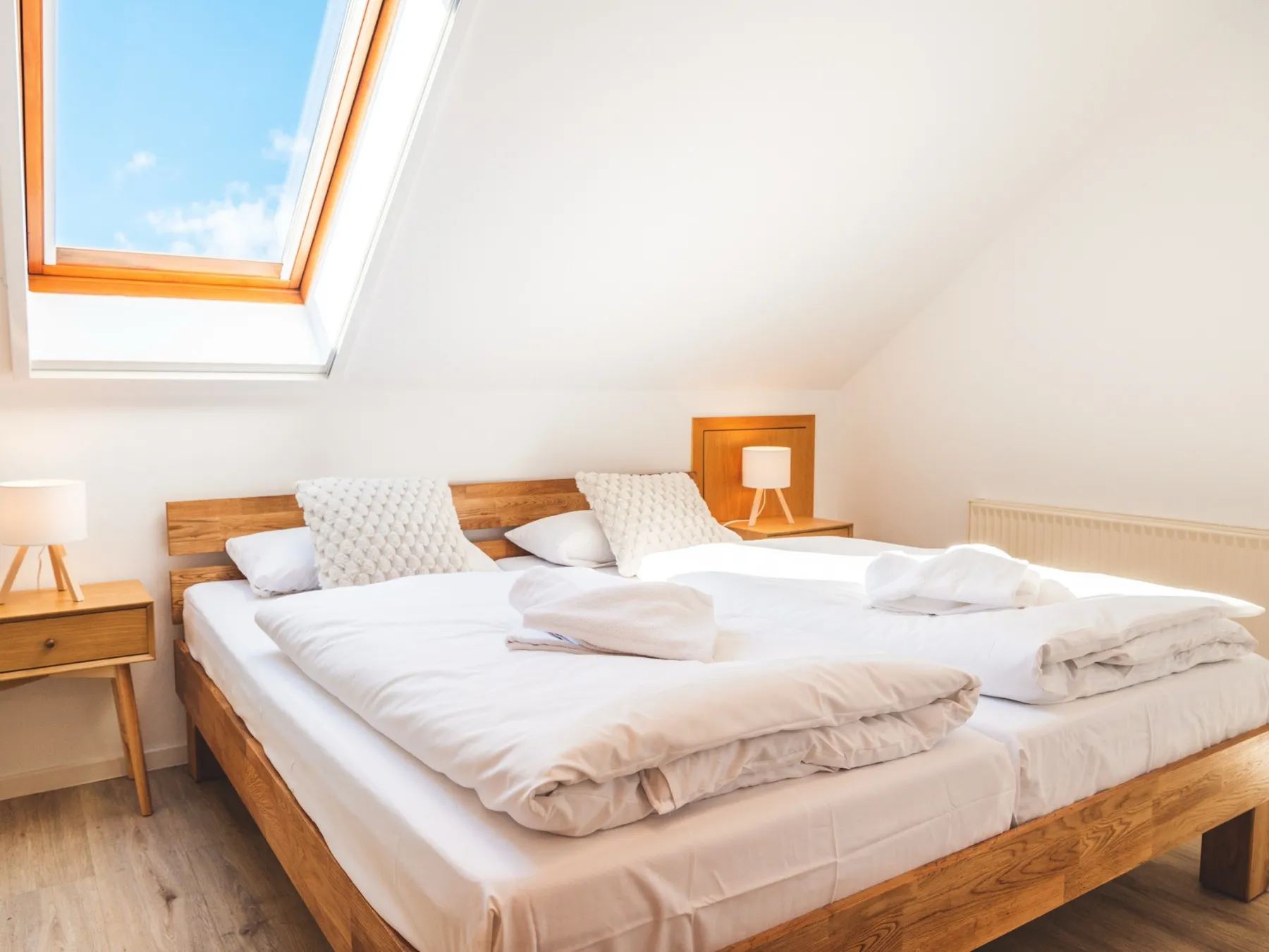 Das Schlafzimmer unter der Dachschräge: Doppelbett mit Eichenrahmen und hölzernem Kopfteil, links oben das Dachfenster mit blauem Himmel und einer weißen Wolke, links und rechts je ein Nachttisch aus Eiche mit Dreibein-Leuchte, auf dem frisch bezogenen Bett zusammengelegte Handtücher