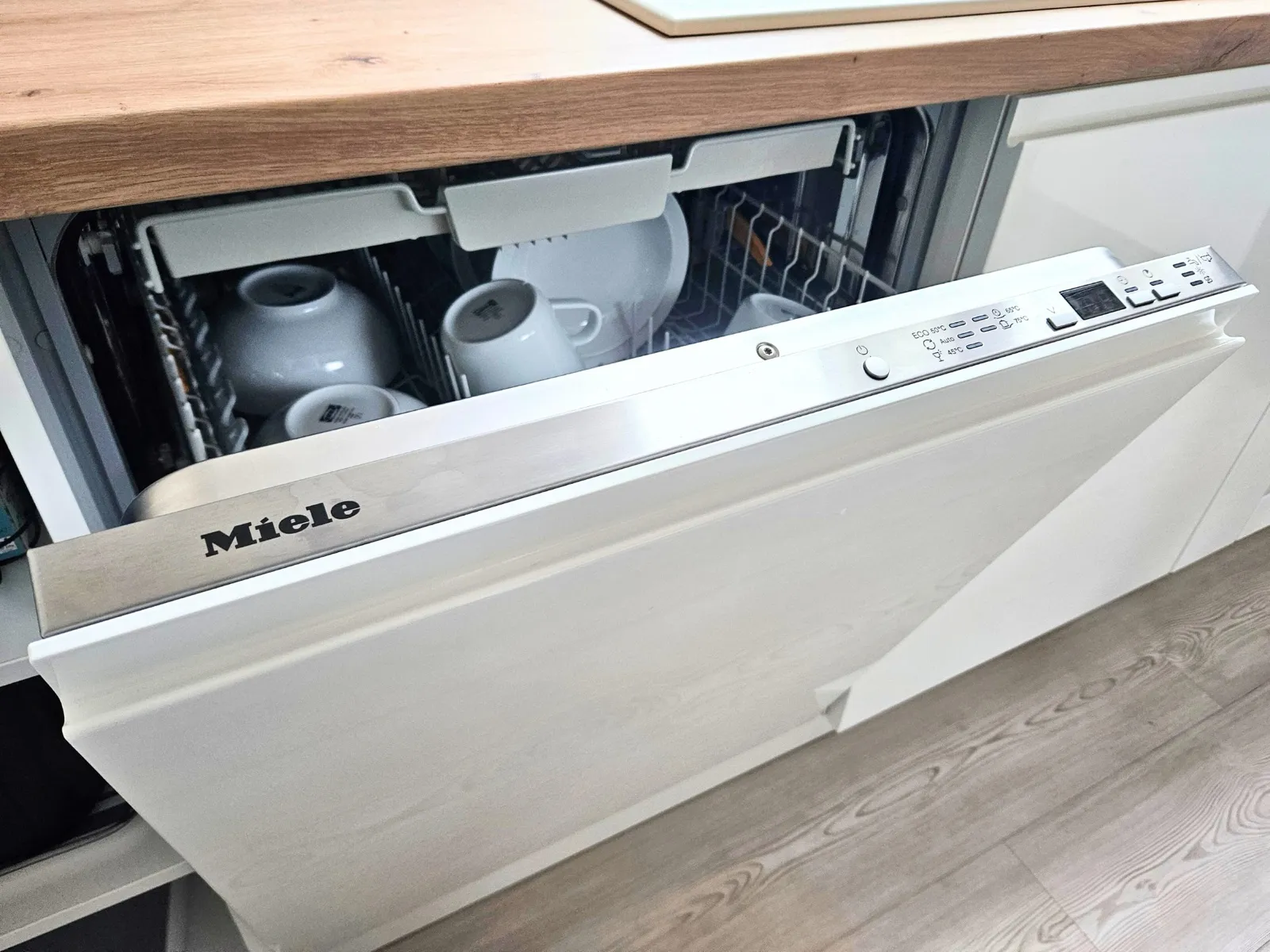 Halb geöffnete, in die weiße Küchenfront integrierte Miele-Spülmaschine unter der hellen Holzarbeitsplatte, im Oberkorb weiße Tassen und Schalen