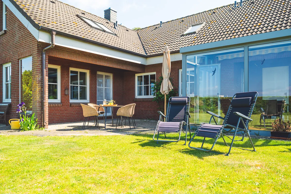 Die eigene Terrasse an der Hausecke: Korbstühle um einen runden Tisch auf Kies, daneben ein Sonnenschirm und zwei dunkelblaue Relaxliegen auf dem Rasen, rechts der verglaste Wintergarten