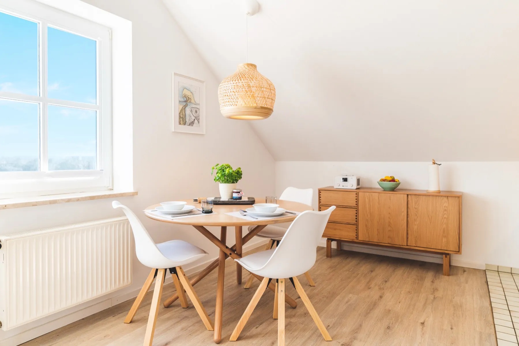Der Essbereich am Fenster: runder Holztisch mit weißen Schalenstühlen, gedeckt für zwei, darüber eine Rattan-Pendelleuchte, rechts ein Sideboard aus Eiche mit Radio und Obstschale, links das Fenster über dem Heizkörper
