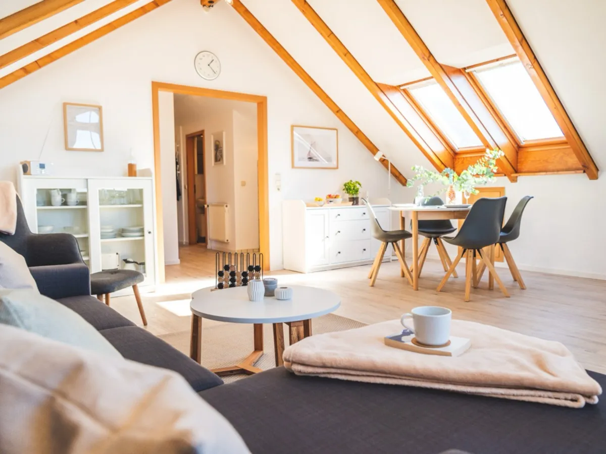 Blick von der Sofaecke über den runden Couchtisch zum Esstisch mit vier dunklen Schalenstühlen unter zwei großen Dachflächenfenstern; links ein weißer Vitrinenschrank mit Geschirr, daneben der Durchgang in den Flur, an der Wand eine runde Uhr, im Vordergrund ein Tablett mit Kaffeetasse auf dem Sofa.