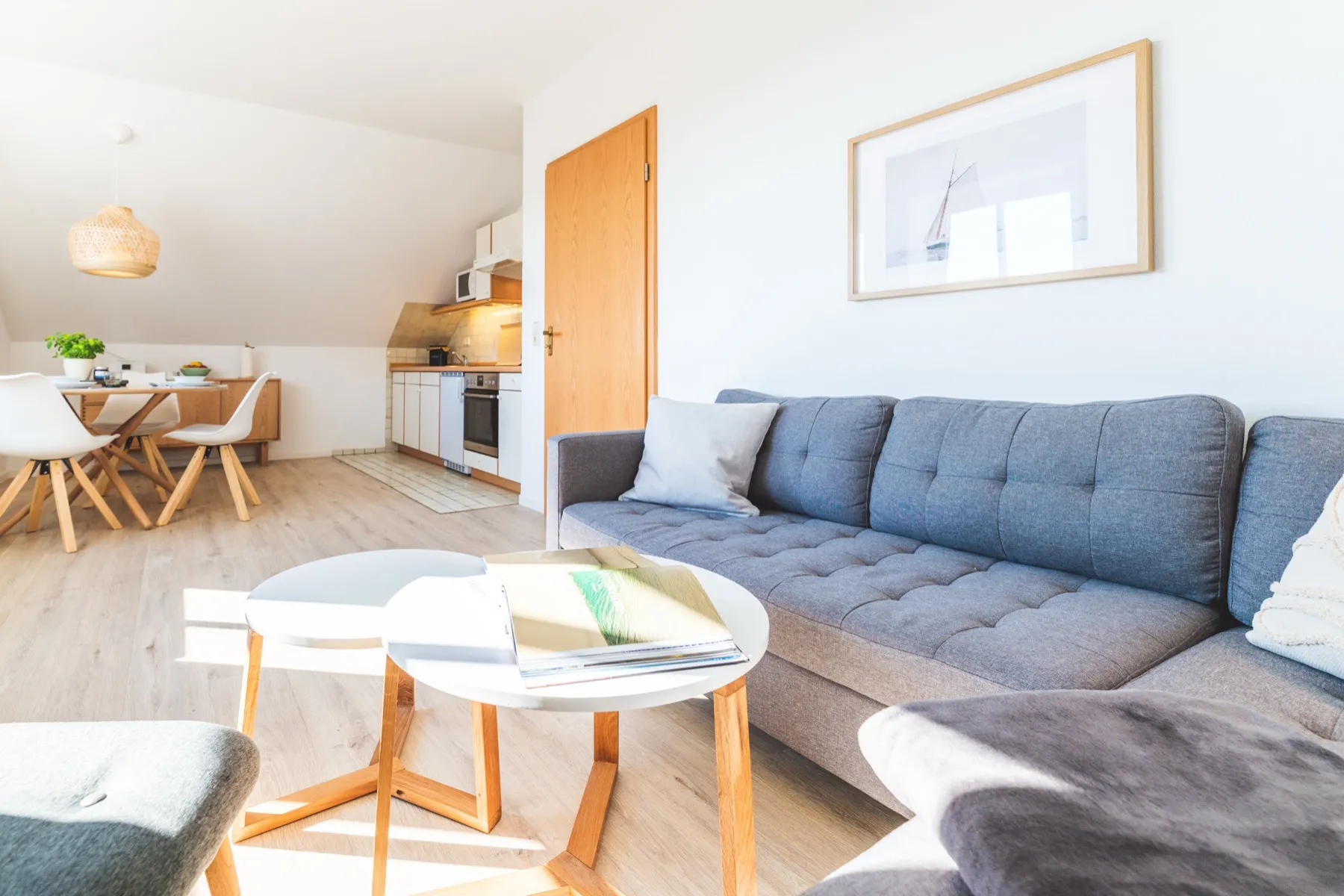 Der Wohnbereich der OG 1 in der Gesamtansicht: langes graues Sofa mit Kissen und Decke, davor zwei runde Beistelltische mit weißer Platte auf Holzbeinen, an der Wand ein gerahmtes Segelboot-Bild; links der runde Esstisch unter der Rattanlampe und dahinter die Küchenzeile