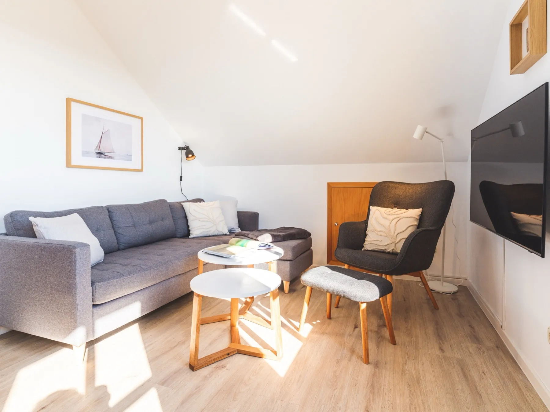 Der Wohnbereich von der Fensterseite aus: das graue Sofa mit Kissen und angebautem Hocker unter der Dachschräge, darüber ein gerahmtes Segelschiff-Bild und eine Wandleuchte, davor zwei runde Beistelltische mit weißer Platte, rechts der dunkle Ohrensessel mit Fußbank, eine Stehleuchte und der Flachbildfernseher an der Wand; Sonnenlicht liegt auf dem Dielenboden