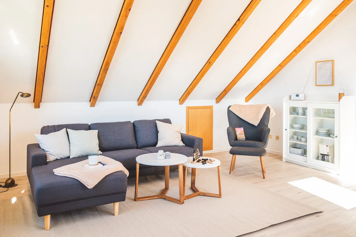 Sitzecke mit dunkelblauem Sofa, Kissen und Wolldecke, davor zwei runde Beistelltische, rechts ein Geschirrschrank mit Glastüren
