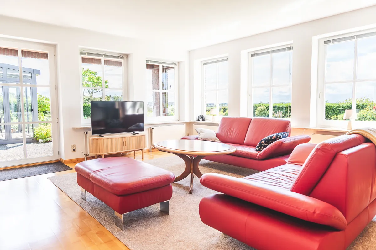 Rote Ledersofas um einen ovalen Couchtisch, davor ein Smart-TV auf einem Sideboard, ringsum Fenster mit Blick ins Grüne
