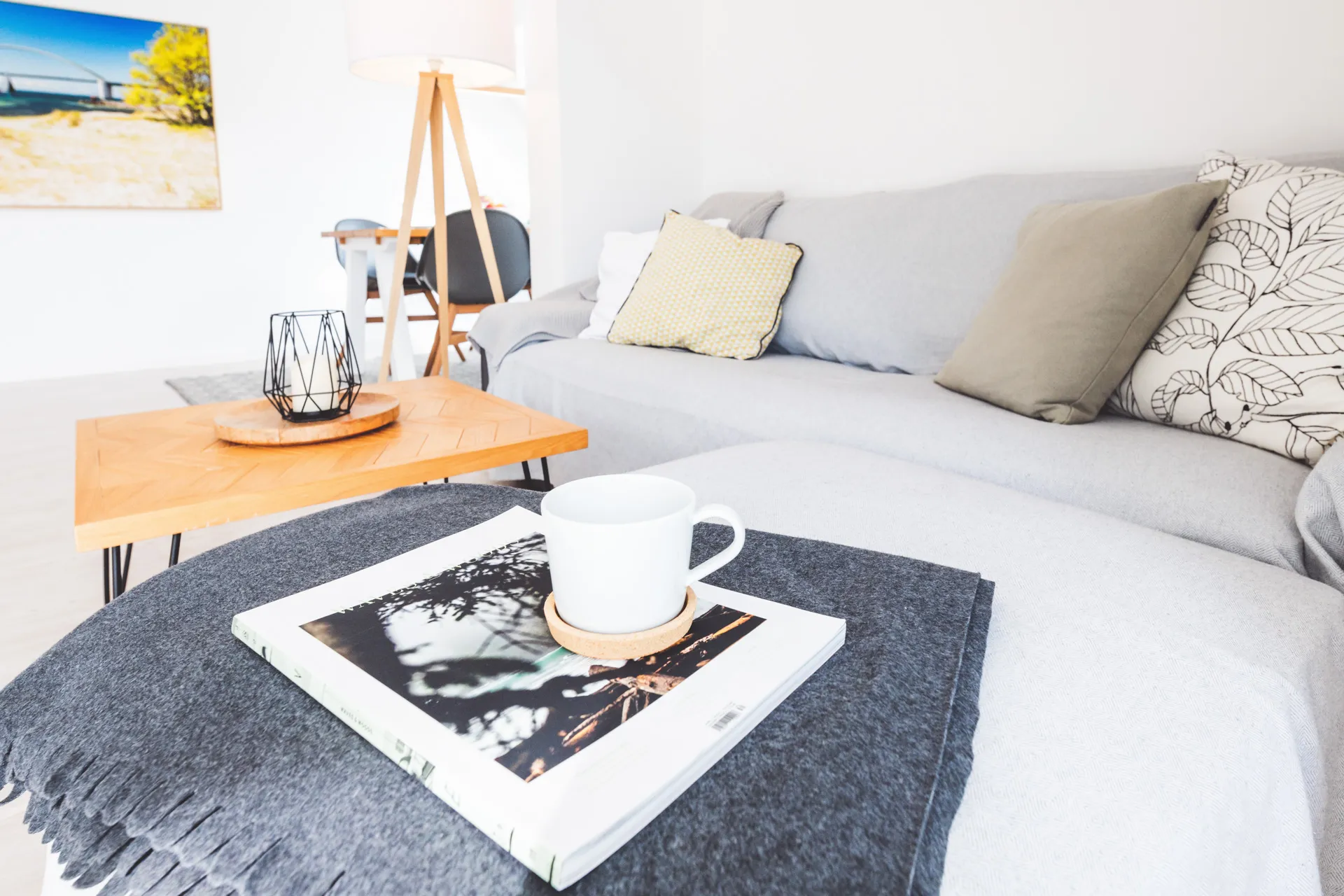 Wohnbereich mit hellgrauem Sofa, Kissen und Wolldecke, davor ein Couchtisch aus Holz; im Vordergrund eine Tasse auf einem Bildband, im Hintergrund der Essplatz