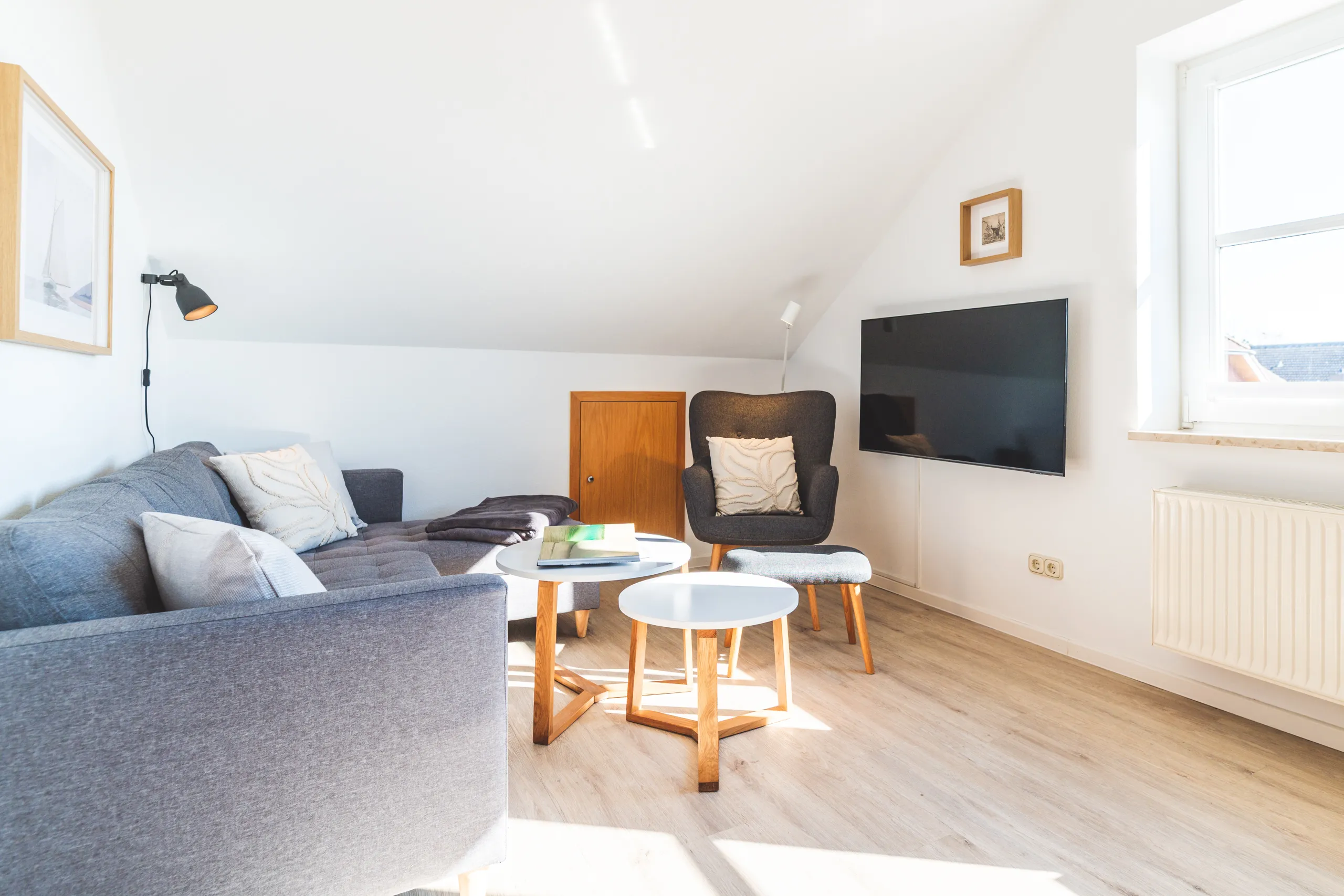 Wohnbereich unter der Dachschräge mit grauem Sofa, dunklem Ohrensessel mit Hocker, zwei runden Beistelltischen und wandhängendem Smart-TV